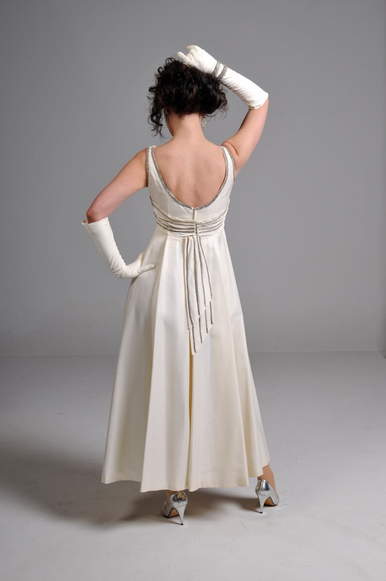 May include: A white floor-length gown with a low back and a beaded detail down the back. The gown is worn by a person with dark hair and white gloves. The person is wearing silver heels.