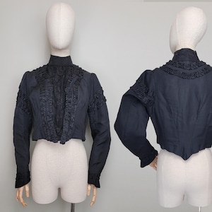 May include: A black cropped jacket with long sleeves and a high collar. The jacket features decorative ruffles along the front and sleeves. The garment is displayed on a mannequin.