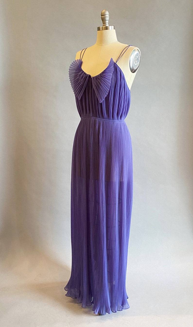 May include: A long, purple, pleated dress with a bow at the neckline. The dress is sleeveless and has a flowy, feminine silhouette.