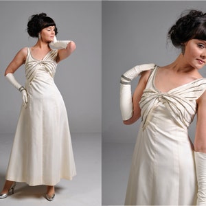 May include: A white sleeveless gown with a beaded bow detail at the neckline. The gown has a flowing skirt and is worn with white gloves.