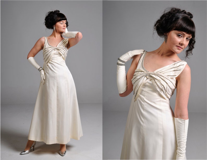 May include: A white sleeveless gown with a beaded bow detail at the neckline. The gown has a flowing skirt and is worn with white gloves.