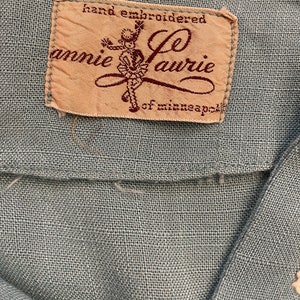 May include: A close-up of a beige fabric label with brown text that reads "hand embroidered Annie Laurie of Minneapolis". The label features a stylized illustration of a girl in a dress.
