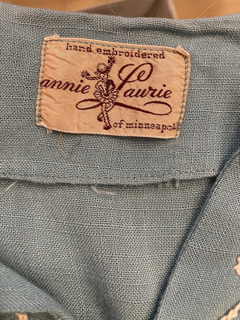 May include: A close-up of a beige fabric label with brown text that reads "hand embroidered Annie Laurie of Minneapolis". The label features a stylized illustration of a girl in a dress.
