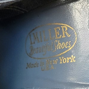 K&ouml;nnte beinhalten: Ein goldfarbenes ovales Etikett mit dem Text "Miller Beautiful Shoes" und "Made in New York U.S.A." auf blauem Hintergrund.