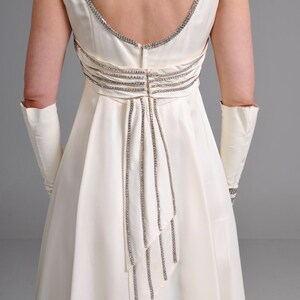 May include: A white satin gown with a low back and a beaded design. The gown has a fitted bodice and a flowing skirt.