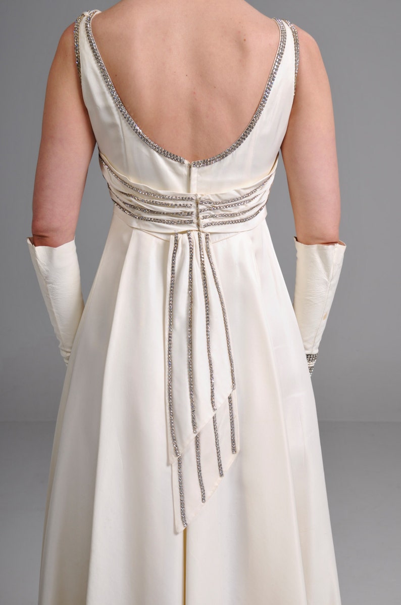 May include: A white satin gown with a low back and a beaded design. The gown has a fitted bodice and a flowing skirt.