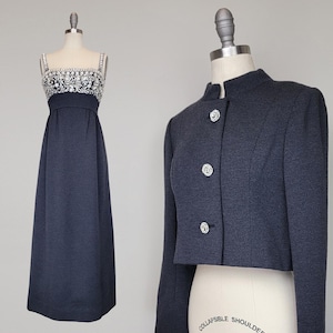 Puede incluir: Una chaqueta corta de manga larga de color gris oscuro con botones plateados y cuello mandarín. La chaqueta se combina con un vestido largo de color gris oscuro con un corpiño de cuentas y pedrería.