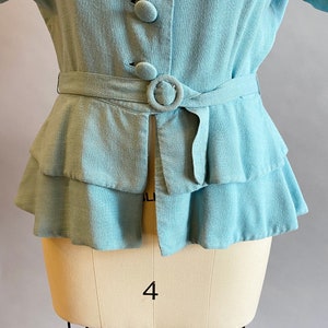 May include: A light blue linen blouse with a ruffled hem and a belt. The blouse has a button-up front and a collar.