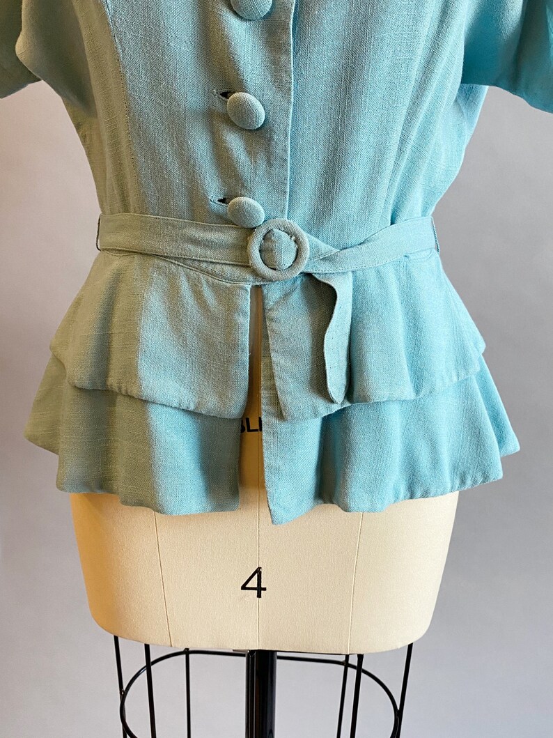 May include: A light blue linen blouse with a ruffled hem and a belt. The blouse has a button-up front and a collar.