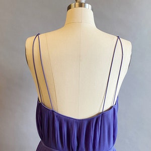 May include: A purple pleated sleeveless dress with thin straps. The dress has a fitted bodice and a flowing skirt.