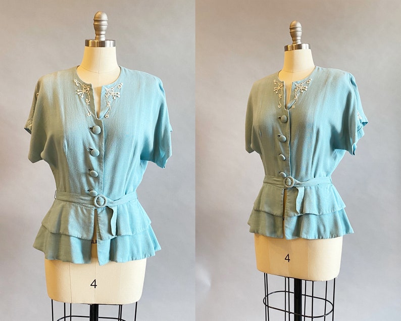 May include: A light blue blouse with a ruffled hem and a beaded neckline. The blouse has short sleeves and a button-up front.