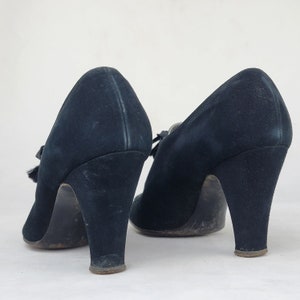 K&ouml;nnte beinhalten: Ein Paar schwarze Wildlederschuhe mit Absatz und einer kleinen Schleife vorne. Die Schuhe haben einen hohen Absatz und eine geschlossene Spitze.