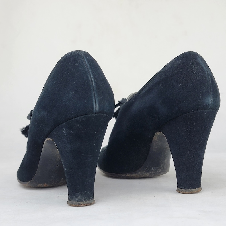 K&ouml;nnte beinhalten: Ein Paar schwarze Wildlederschuhe mit Absatz und einer kleinen Schleife vorne. Die Schuhe haben einen hohen Absatz und eine geschlossene Spitze.