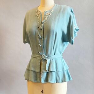 May include: A light blue blouse with a ruffled hem and a belt. The blouse has a V-neckline and short sleeves. The neckline and sleeves are decorated with white embroidery.