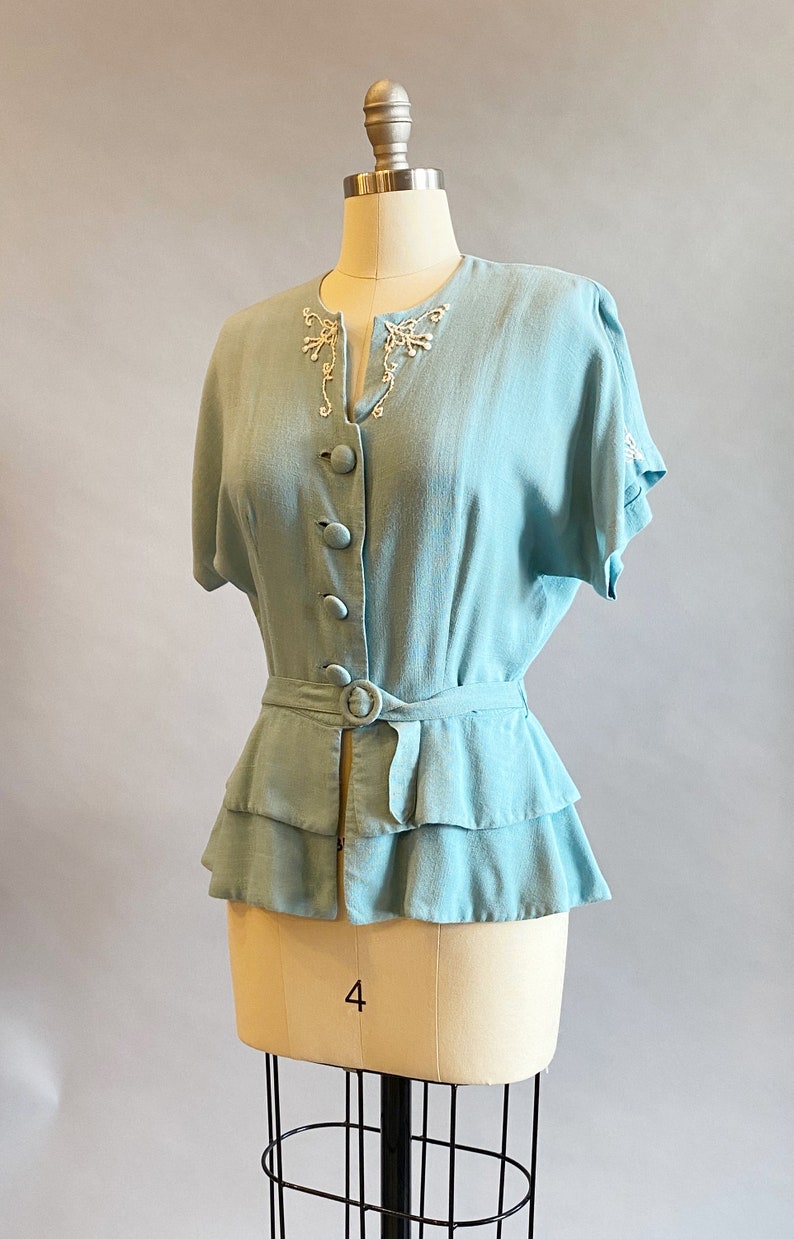 May include: A light blue blouse with a ruffled hem and a belt. The blouse has a V-neckline and short sleeves. The neckline and sleeves are decorated with white embroidery.