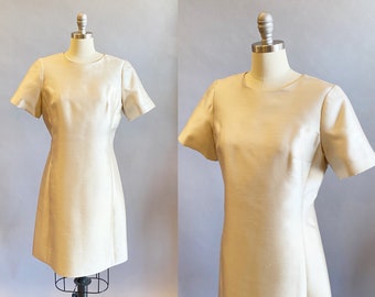 May include: A cream-colored, short-sleeved dress displayed on a mannequin. The dress has a simple, elegant design with a round neckline and a slightly flared skirt. The fabric appears to have a subtle sheen, suggesting a silk or satin material.