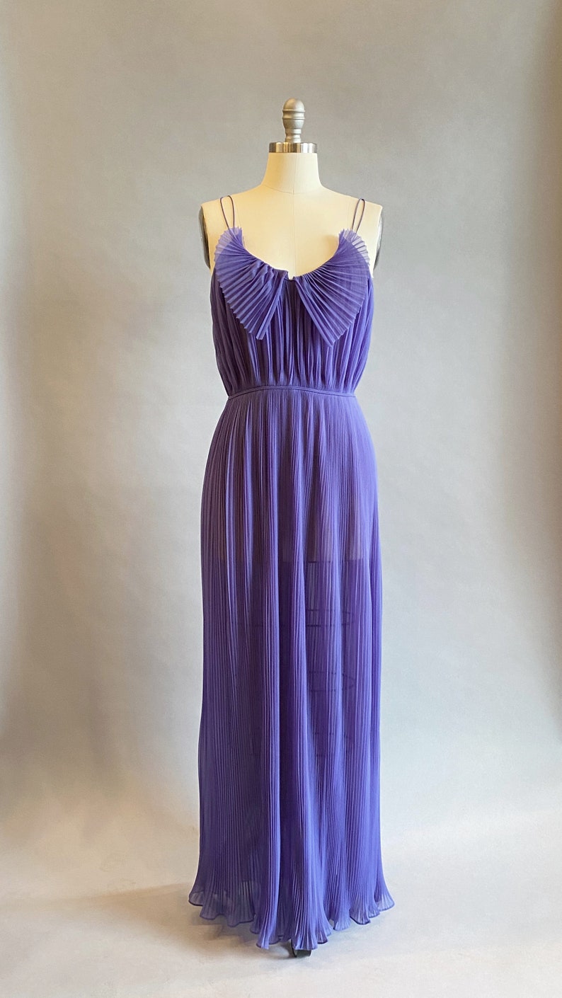 May include: A long, purple, pleated maxi dress with thin straps and a ruffled neckline. The dress is on a white mannequin against a light gray background.