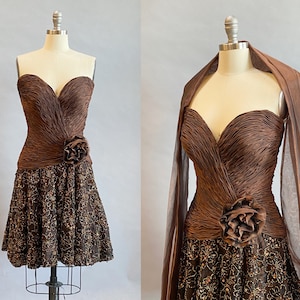 Op de afbeelding: Een bruine strapless jurk met een gerimpelde buste en een rok met pailletten. De jurk heeft een grote bruine bloem als accent op de taille.