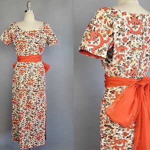 May include: A white dress with a red and orange floral print featuring dragons. The dress has a fitted bodice and a full skirt. It is accessorized with a matching red sash and a sheer red scarf.