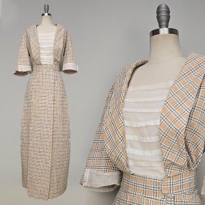May include: A vintage-style ensemble on a mannequin. The outfit includes a long, plaid skirt and a matching jacket with rolled-up sleeves. A white, layered top is visible beneath the jacket. The plaid pattern features tan, blue, and white lines.