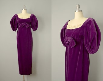 May include: A long purple velvet dress with puffed sleeves and a bow at the waist. The dress has a square neckline and a fitted bodice.
