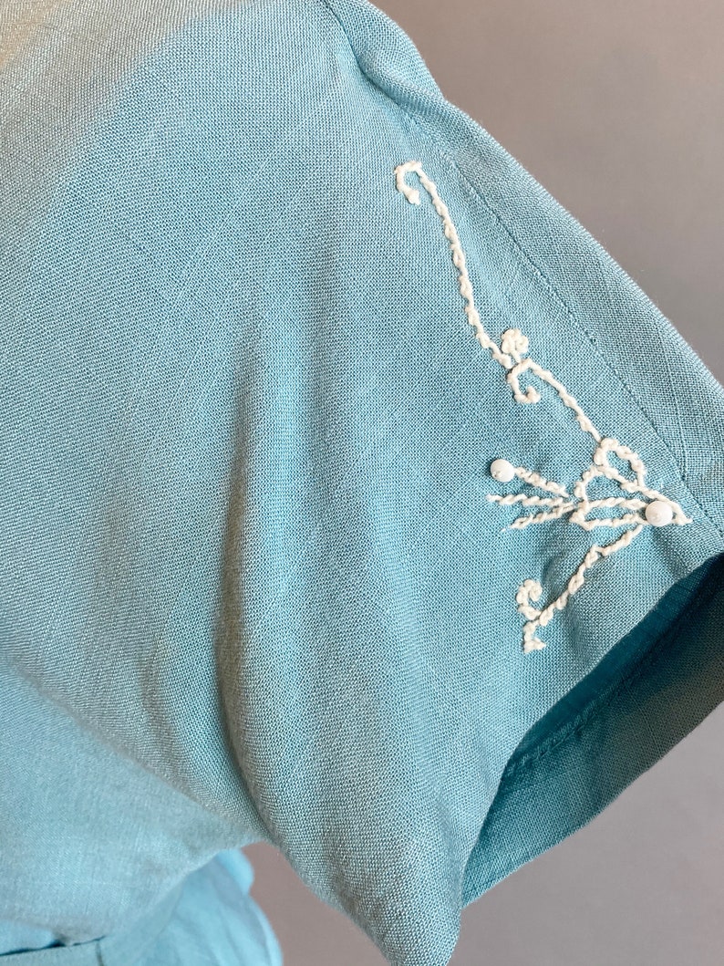 May include: A light blue linen dress with white embroidered details on the sleeve.