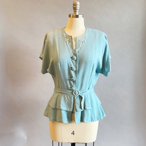 May include: A light blue blouse with a ruffled hem and a white embroidered neckline. The blouse has a belt and buttons down the front.