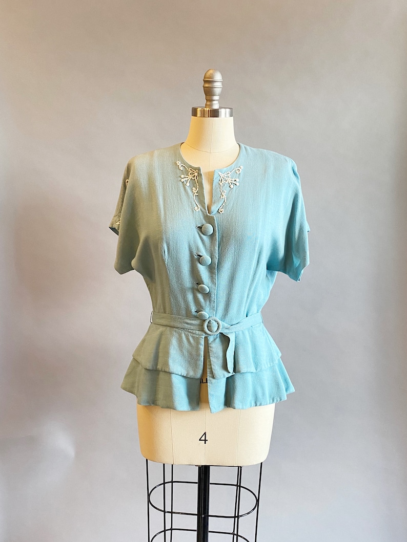 May include: A light blue blouse with a ruffled hem and a white embroidered neckline. The blouse has a belt and buttons down the front.