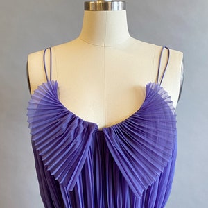 May include: A purple pleated sleeveless dress with a ruffled neckline. The dress is made of a sheer fabric and has a fitted bodice.