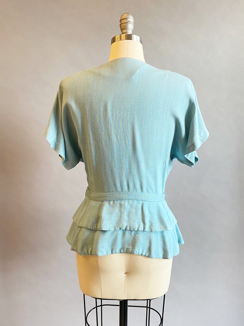 May include: A light blue blouse with a ruffled hem and a tie at the waist.