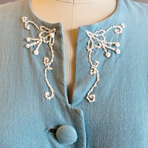 May include: A light blue linen jacket with white embroidered details around the neckline and a single button closure.