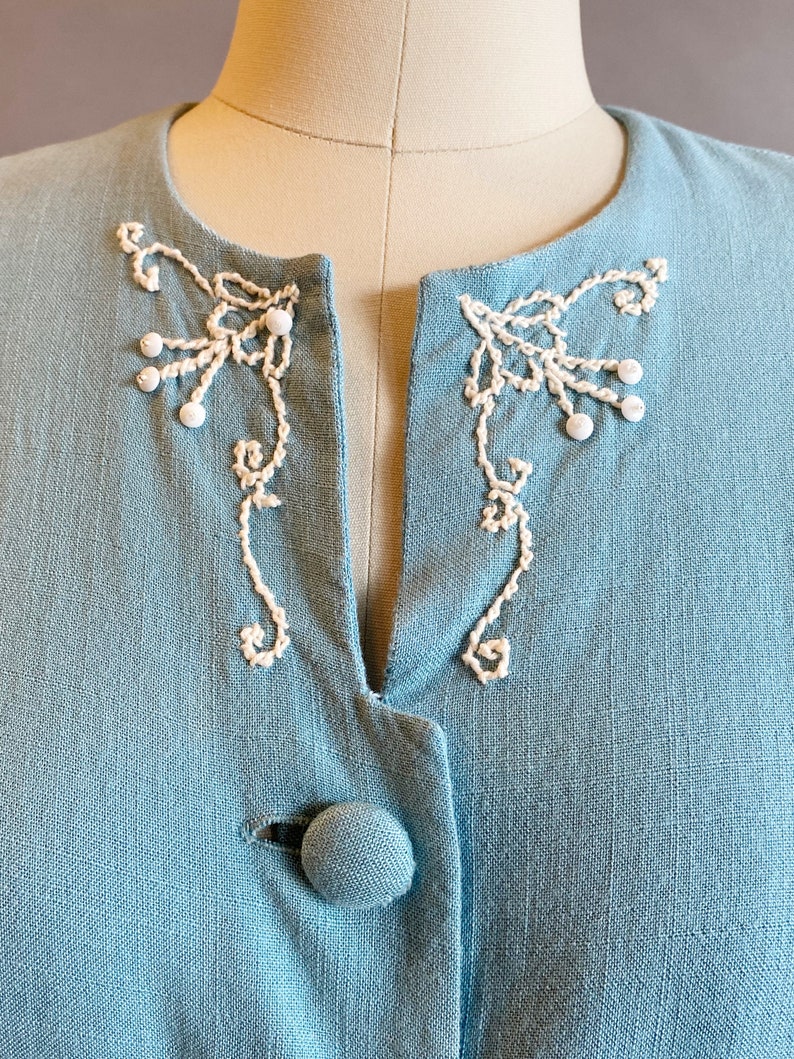 May include: A light blue linen jacket with white embroidered details around the neckline and a single button closure.