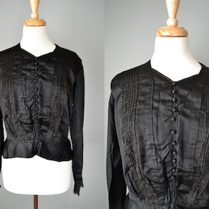May include: A black satin blouse with a ruffled hem and long sleeves. The blouse has a button-up front and is decorated with gold stitching.
