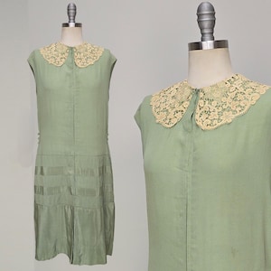 May include: A pale green vintage dress with a cream lace collar. The sleeveless dress has a pleated skirt and a button-up front. The dress is displayed on a mannequin.