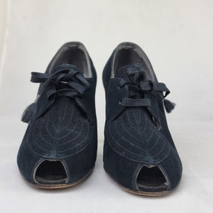 K&ouml;nnte beinhalten: Ein Paar schwarze Wildlederschuhe mit Peeptoe, Schn&uuml;rung und Quasten. Die Schuhe haben einen hohen Absatz und eine spitze Zehenpartie.