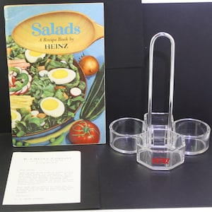May include: A vintage Heinz cookbook titled "Salads: A Recipe Book by Heinz" with a colorful illustration of a salad on the cover.  The cookbook is next to a clear plastic condiment holder with a handle and the Heinz logo.