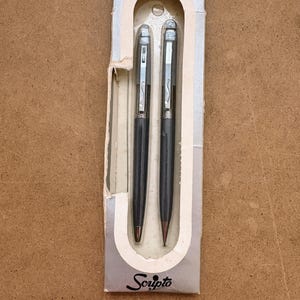 May include: A vintage gift set of two Scripto pens. The pens are dark gray with silver accents and a silver clip. The packaging is a light gray cardboard with the words "Gift Set" and "Scripto" printed on it.
