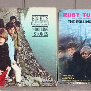 May include: Two vintage vinyl record covers featuring The Rolling Stones. The left cover is blue and green with the text "Big Hits (High Tide and Green Grass)" and "The Rolling Stones". The right cover is blue and white with the text "Ruby Tuesday" and "The Rolling Stones".