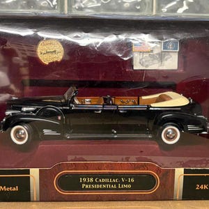 Puede incluir: Un modelo de coche de fundición a presión negro de 1938 Cadillac V-16 Presidential Limo en una caja de exhibición. La caja tiene una moneda chapada en oro y el texto "Scale 1:24", "Die Cast Metal", "1938 Cadillac V-16 Presidential Limo" y "24K Gold Plated Coin".