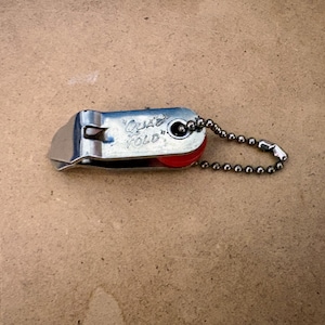 May include: A small, silver-colored utility tool with a red roller and a ball chain. The text "Quad Fold" is visible on the tool. The tool is designed to fold and is likely used for cutting or trimming.