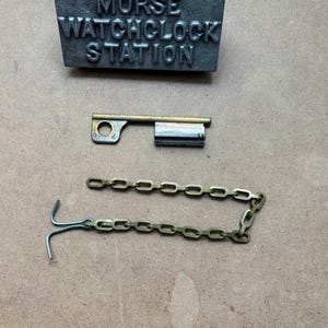 May include: A collection of antique watch clock parts. The set includes a metal plate with the text "MORSE WATCHCLOCK STATION", a key, and a chain with a hook. The items are arranged on a light brown surface.