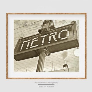 May include: Sepia-toned framed print of a vintage Metro sign. The sign, with the word "METRO" in bold white letters, is mounted on a fluted column. The image is framed in a light brown wooden frame.