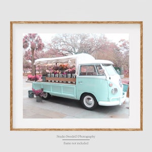 May include: A light blue vintage van with a white roof and white trim. The van is parked on a street with a pink and green background. The van is filled with flowers in pots. The van has a white awning over the back. The text 
