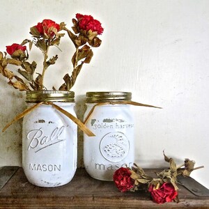 May include: Two white painted mason jars with gold lids and brown twine. The jars are filled with dried red flowers. The jars are sitting on a wooden surface. The text on the jars reads "Ball Mason" and "golden harvest mason".