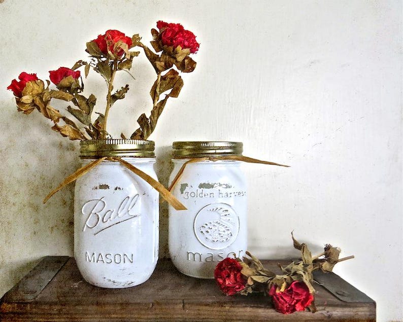 May include: Two white painted mason jars with gold lids and brown twine. The jars are filled with dried red flowers. The jars are sitting on a wooden surface. The text on the jars reads "Ball Mason" and "golden harvest mason".
