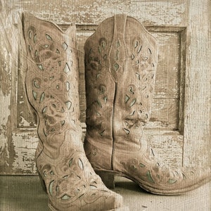 Op de afbeelding: Een paar bruine leren cowboylaarzen met ingewikkelde uitgesneden ontwerpen. De laarzen staan tegen een rustieke houten achtergrond.