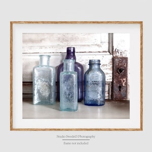 May include: Four vintage glass bottles in shades of blue and purple, arranged on a whitewashed wooden surface. The bottles have a frosted, aged appearance and are grouped together in a rustic setting.