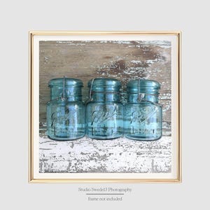 May include: Three blue glass jars with metal lids and wire handles sit on a weathered wooden surface. The jars are arranged in a row, with the middle jar slightly higher than the others. The background is a light blue and white wood grain.