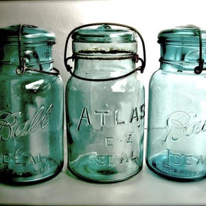 May include: Three vintage blue-green glass Ball and Atlas E-Z Seal canning jars. The jars have metal lids and wire closures. The Ball jars have "Ball Ideal" embossed on the front. The Atlas jar has "Atlas E-Z Seal" embossed on the front.