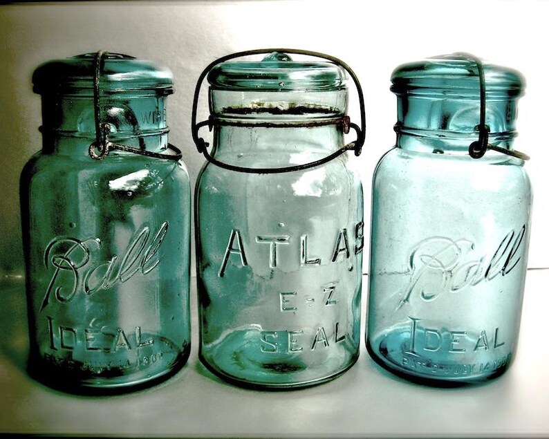 May include: Three vintage blue-green glass Ball and Atlas E-Z Seal canning jars. The jars have metal lids and wire closures. The Ball jars have "Ball Ideal" embossed on the front. The Atlas jar has "Atlas E-Z Seal" embossed on the front.